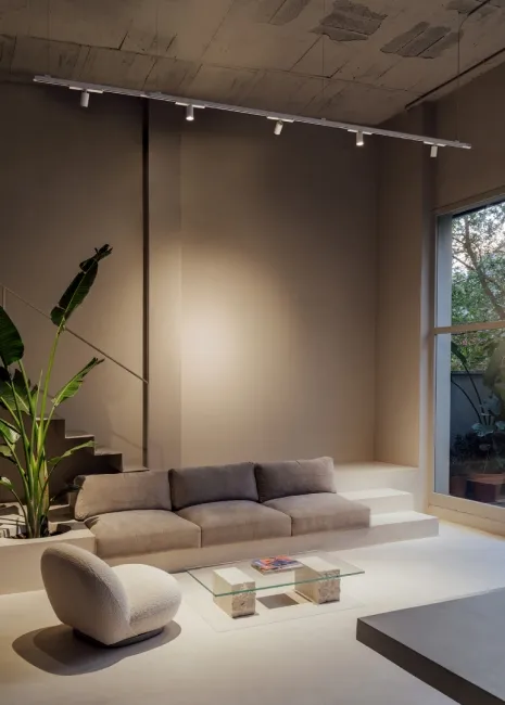 Modern living room with beige sofa, coffee table, and track lighting, next to a large window with plants.