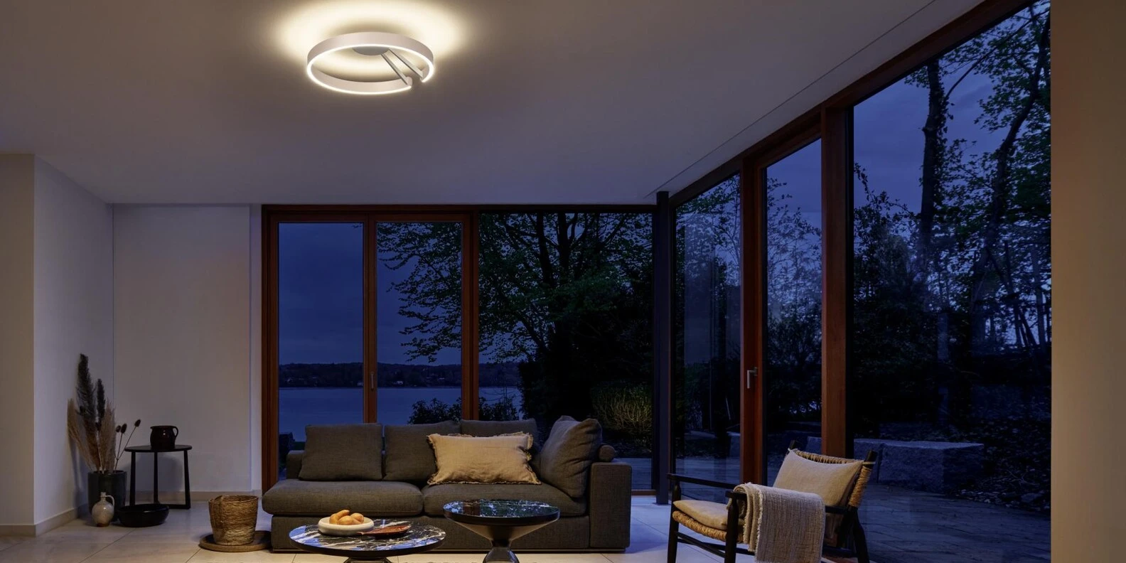 Modern living room at dusk with large windows overlooking a forest and lake.
